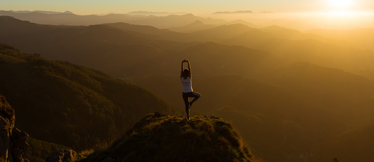 Silhouette of a person in a yoga tree pose on a hilltop overlooking misty mountains at sunset, warm golden tones